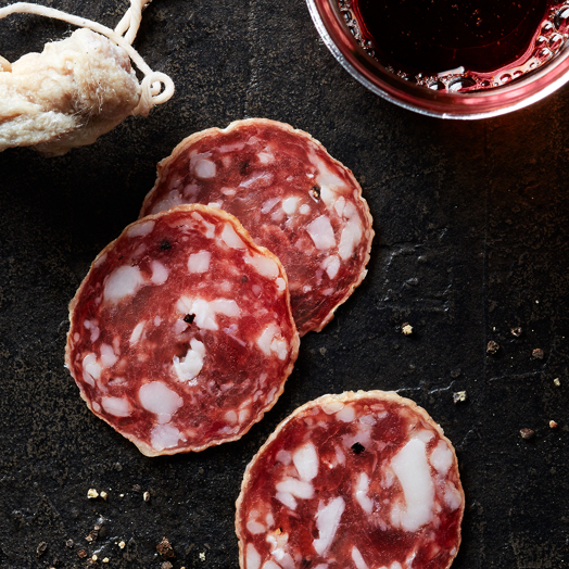 Three slices of Columbus Salami on a black slate sprinkled with black peppercorns and a glass of red wine